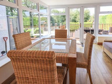 A sunroom with a glass top table and four wicker chairs overlooking a wooden deck at Ellerbeck Bridge in Underbarrow near Kendal