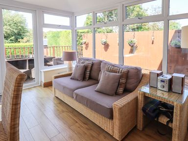 A sunroom with a wicker sofa and cushions a wicker chair a side table with a lamp and stereo and windows overlooking an outdoor seating area at Ellerbeck Bridge in Underbarrow near Kendal
