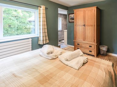 A bedroom with a bed topped with folded towels a wooden wardrobe a trash bin and a window with checked curtains at Ellerbeck Bridge in Underbarrow near Kendal