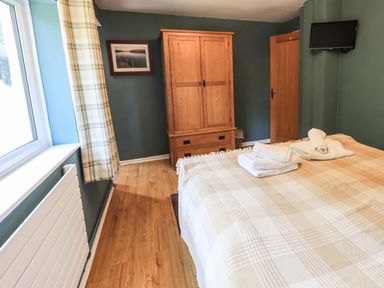A bedroom with a wooden wardrobe a bed with folded towels a wall-mounted TV and a large window with checkered curtains at Ellerbeck Bridge in Underbarrow near Kendal