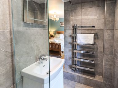A bathroom with a white sink and chrome towel rack with a white towel and a glimpse of a bedroom with a wooden bed frame and lamp at Ellerbeck Bridge in Underbarrow near Kendal
