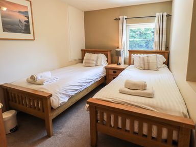 A bedroom with two single beds with white linens and towels a bedside table with a lamp and a window with curtains at Ellerbeck Bridge in Underbarrow near Kendal