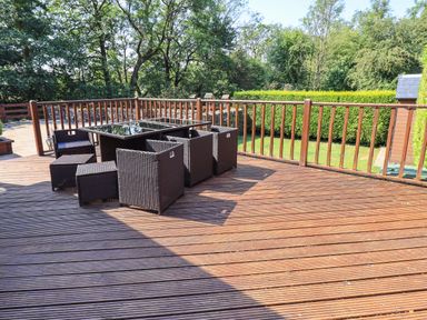 An outdoor wooden deck with a glass top table and wicker chairs surrounded by trees and hedges at Ellerbeck Bridge in Underbarrow near Kendal