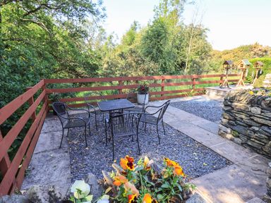 An outdoor patio area with a metal table and chairs surrounded by trees and flowers at Ellerbeck Bridge in Underbarrow near Kendal