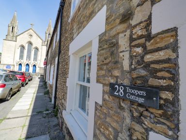 Stone building with a sign reading 28 Troopers Cottage next to a sidewalk with parked cars and a church in the background in Frome