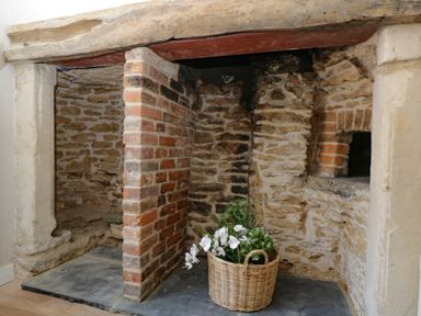 A stone and brick fireplace with a wooden beam on top and a basket with white flowers inside at Troopers Cottage in Frome