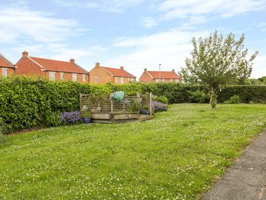A garden with grass a tree flower pots and a wooden deck with houses and hedges in the background at Sea-u-Soon in Runswick Bay