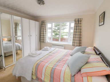 A bedroom with a bed covered in patterned bedding next to a window with checkered curtains and white wardrobes with mirrors at Sea-u-Soon in Runswick Bay