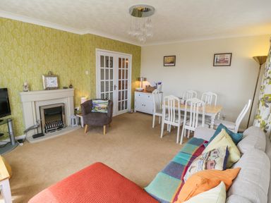 A living room with a sofa covered in colorful cushions and a blanket a dining table with six chairs a small armchair next to a fireplace and a cabinet at Sea-u-Soon in Runswick Bay