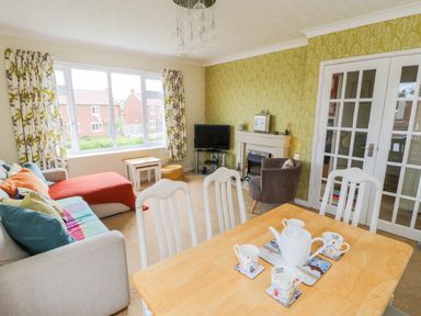 A living room with a sofa, armchair, small tables, TV, fireplace, dining table with teapot and cups at Sea-u-Soon in Runswick Bay