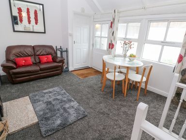 A living room with a brown leather sofa with red cushions a round dining table with four chairs a vase of flowers and a bowl of fruit near windows at Toadstools in Rame Cross near Penryn