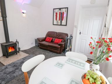 A room with a round white table with chairs a fruit bowl and flower vase a brown leather sofa and a wood burning stove at Toadstools in Rame Cross near Penryn
