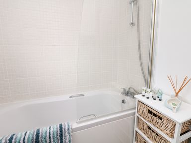 A bathroom with a bathtub and shower alongside a small cabinet with wicker baskets and toiletries at Quayside View in Brixham