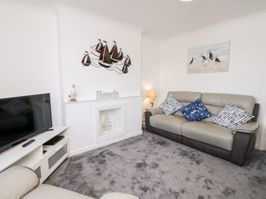 A living room with a TV on a white stand a gray sofa three cushions a lamp and wall decorations at Quayside View in Brixham