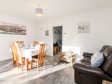 A dining room with wooden chairs around a table next to a brown leather sofa and wicker table at Quayside View in Brixham