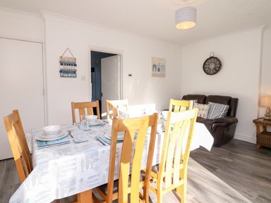 A dining room with a wooden table set with dishes and glasses and a black leather sofa with cushions in a living area at Quayside View in Brixham