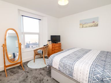 A bedroom with a bed covered in patterned bedding a wooden full-length mirror a wicker chair on a rug and a wooden chest of drawers with a TV on top at Quayside View in Brixham