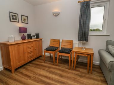 A room with a wooden sideboard, two wooden chairs with cushions, nested wooden tables, and a window at 64 Waterside in Lanivet
