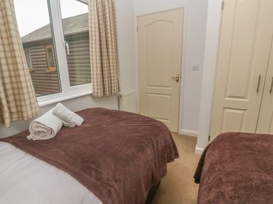 A bedroom with two beds covered in brown blankets a window with checkered curtains a radiator a door and a built in wardrobe at 64 Waterside in Lanivet