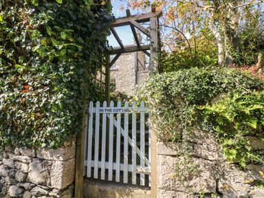 A garden gate leading to a stone house with greenery and a wooden arbor at Westmorland Cottage in Grange-Over-Sands