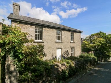 A stone house with white windows and a white door surrounded by greenery next to a road at Westmorland Cottage Grange-Over-Sands