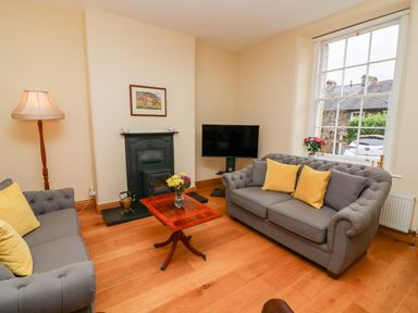 A living room with two grey sofas yellow cushions a wooden coffee table a fireplace a floor lamp a television and a large window at Westmorland Cottage in Grange-Over-Sands