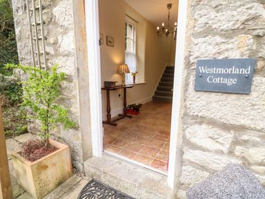 The entrance to a stone cottage showing a tiled hallway with a small table lamp and a staircase at Westmorland Cottage Grange-Over-Sands