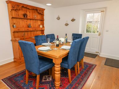 A dining room with a wooden table set with plates glasses and bottles surrounded by blue chairs and a wooden cabinet against the wall at Westmorland Cottage in Grange-Over-Sands
