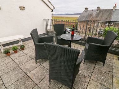 An outdoor patio with four black wicker chairs around a round table with a bottle and two glasses overlooking fields and houses at Westmorland Cottage in Grange Over Sands