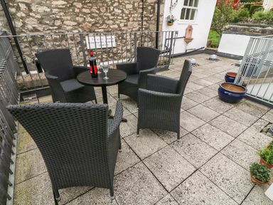 An outdoor patio with four black wicker chairs and a round table with a bottle and two glasses at Westmorland Cottage in Grange-Over-Sands