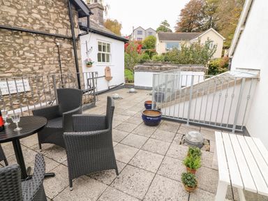 A patio with black wicker chairs a round black table a white bench potted plants and stone and white buildings in the background at Westmorland Cottage in Grange-Over-Sands