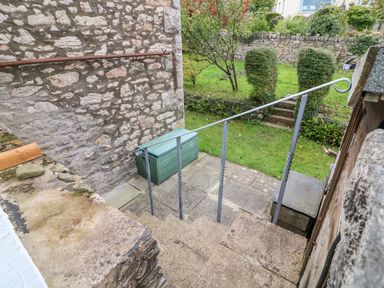 Stone steps with metal handrail leading to a paved area and a green garden shed in a garden with trimmed bushes and stone walls at Westmorland Cottage in Grange-Over-Sands