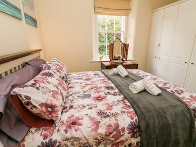 A bedroom with a floral bedspread and rolled towels on the bed a dressing table with a mirror and a window behind it and white wardrobe cabinets at Westmorland Cottage in Grange-Over-Sands