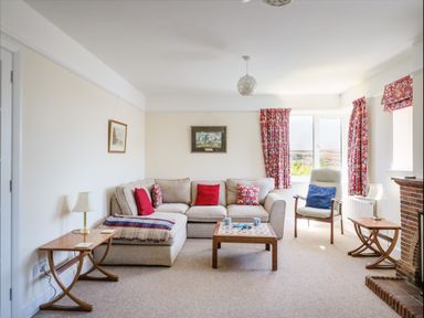 A living room with a beige sofa red and patterned cushions a wooden coffee table armchair with a blue cushion side tables with lamps and red patterned curtains at Thorncroft in West Bay