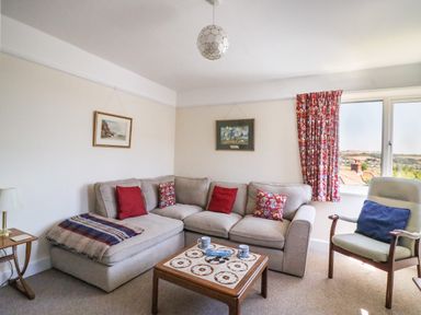 A living room with a beige sofa with red and patterned cushions a patterned throw a wooden table with tiled top with mugs and games a pale green armchair with a blue cushion red patterned curtains a side table with a lamp and two framed pictures on the wall at Thorncroft West Bay