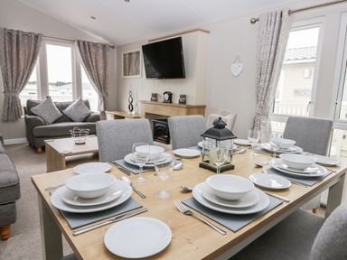 A dining table set for six with white dishes and glasses in a living room with gray sofas and a wall-mounted TV at Cayton Pines Cayton Bay near Scarborough