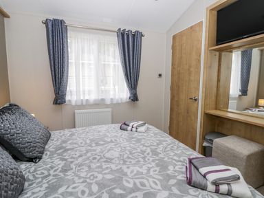 A bedroom with a bed made with patterned gray and white bedding two folded towels on the bed a window with blue patterned curtains a wooden door a wall-mounted TV and a wooden vanity with a mirror and a cushioned stool at Cayton Pines Cayton Bay near Scarborough