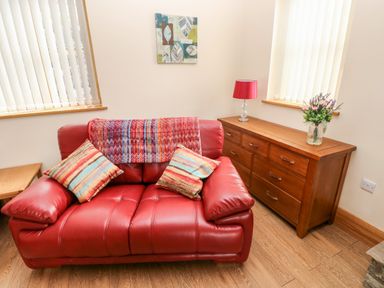 A red leather sofa with two striped cushions and a throw blanket next to a wooden sideboard with a red lamp and a flower arrangement at Grooms Quarters at Pant y Gaseg Farm Ammanford