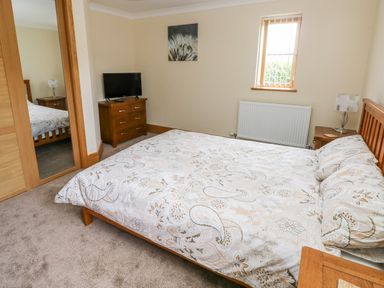 A bedroom with a patterned bedspread bed a wooden dresser with a television a bedside table with a lamp and a window at Grooms Quarters at Pant y Gaseg Farm in Ammanford