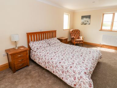 A bedroom with a double bed floral bedding wooden nightstands a lamp an armchair and windows at Grooms Quarters at Pant y Gaseg Farm in Ammanford