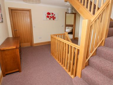 A carpeted hallway with wooden stairs and railing a wooden door a chest of drawers and a bedroom visible through an open door at Grooms Quarters at Pant y Gaseg Farm Ammanford