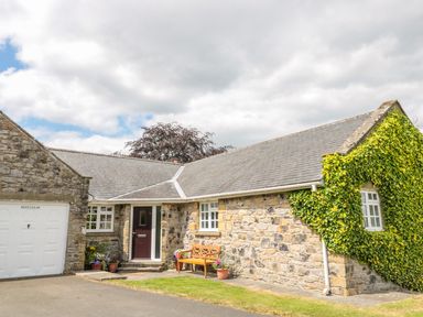 A stone cottage with a single wooden door and white garage door with plants and bench outside at Coquet View Cottage in Rothbury