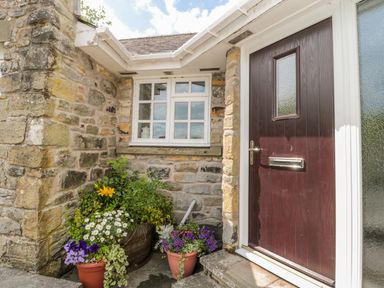 A stone cottage entrance with a dark wooden door white framed window and flower pots at Coquet View Cottage in Rothbury