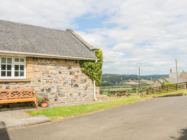 Exterior view of a stone cottage with a wooden bench and potted plant next to a paved driveway and picnic table on a grassy area at Coquet View Cottage in Rothbury