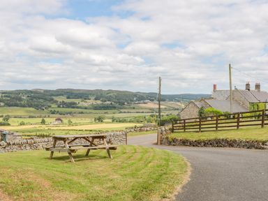 A picnic table on grass near a stone wall and houses with rolling hills and fields in the background at Coquet View Cottage in Rothbury