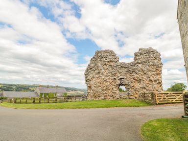 A stone ruin with a small window opening near a wooden fence and gate with houses and hills in the background at Coquet View Cottage in Rothbury