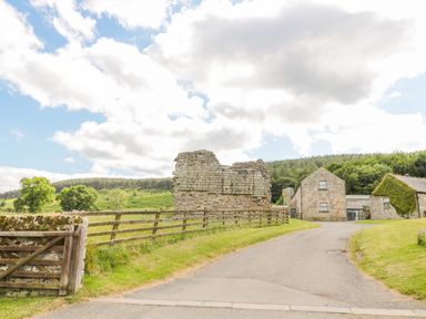 A rural road leading to stone buildings and a stone ruin with wooden fence and green fields surrounding at Coquet View Cottage in Rothbury