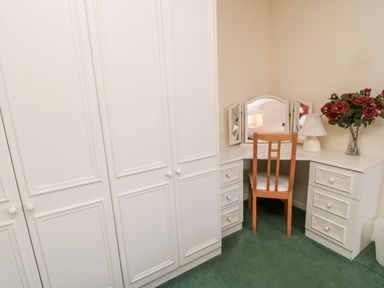 A corner vanity desk with a wooden chair drawers a lamp and a vase with flowers next to a white wardrobe at Coquet View Cottage Rothbury