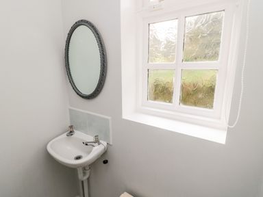 A small bathroom corner with a wall-mounted sink with two taps a round wall mirror and a textured glass window at Coquet View Cottage in Rothbury