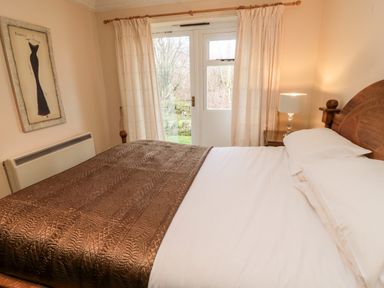 A bedroom with a wooden bed with white sheets and brown blanket a bedside table with lamp and curtains on a glass door at Coquet View Cottage in Rothbury